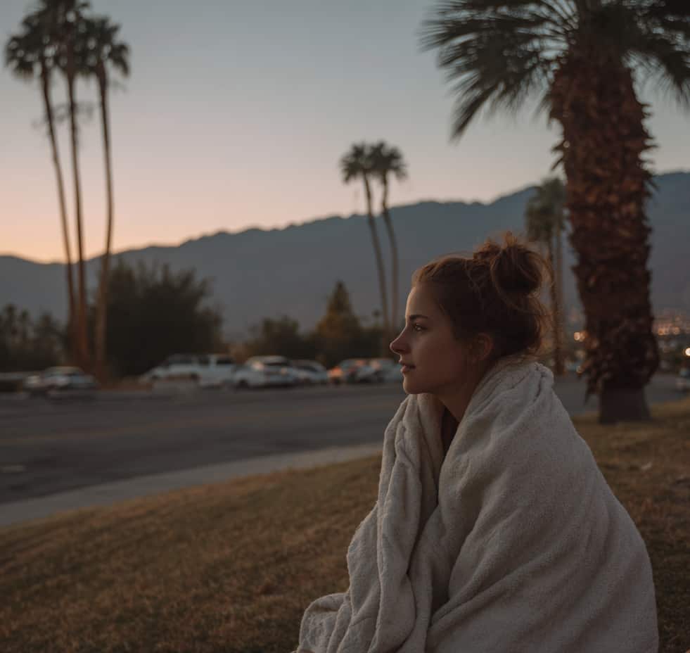 A woman wrapped in a blanket sits peacefully at dusk with palm trees and mountains behind her, evoking the calm of detox in Palm Springs.