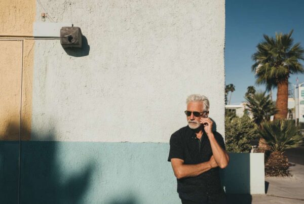 A middle-aged man with gray hair and sunglasses stands against a pastel-colored wall in a palm-lined desert neighborhood, illustrating getting off antidepressants and mental health reflection.