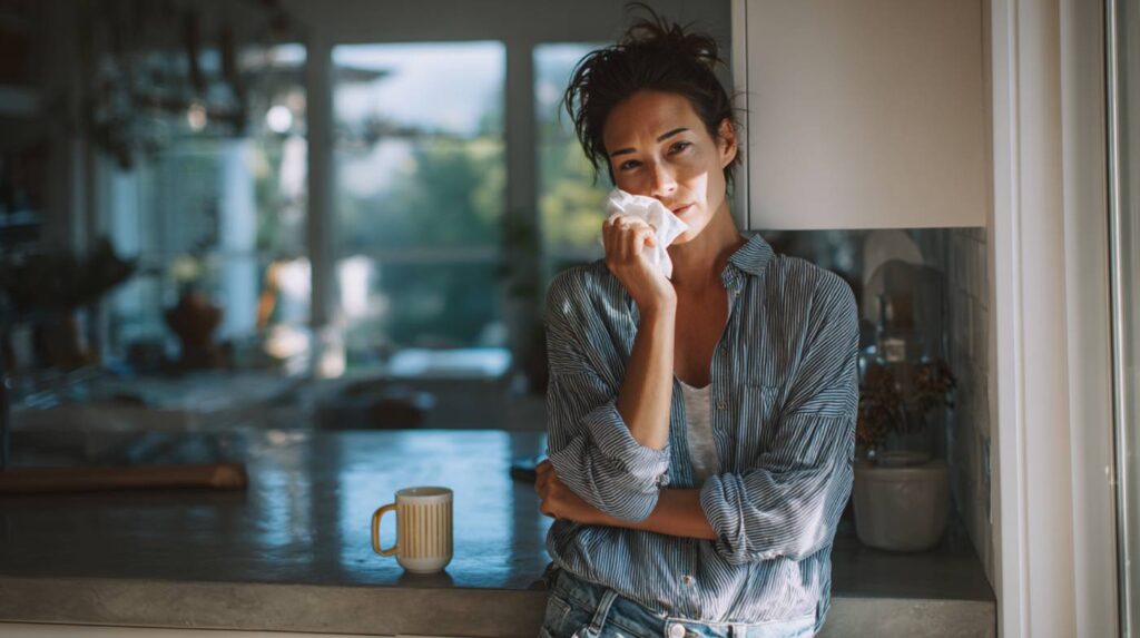 A woman standing at a kitchen counter holding a tissue in soft morning light, illustrating the risks of mixing DayQuil and alcohol.